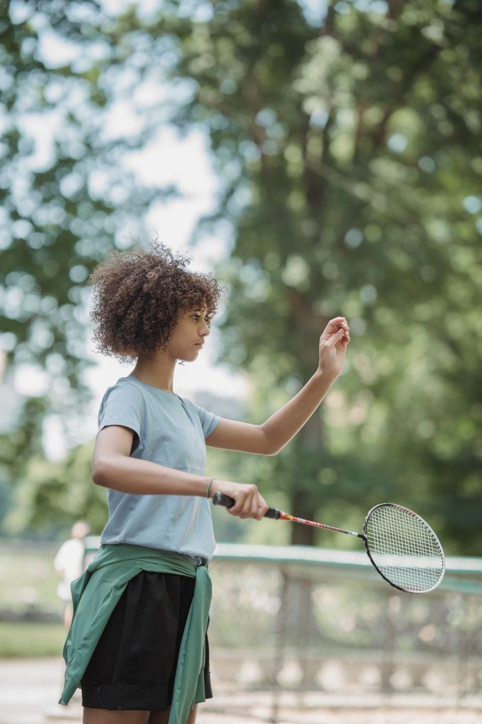 Young girl with curly hair playing badminton in a sunny park.